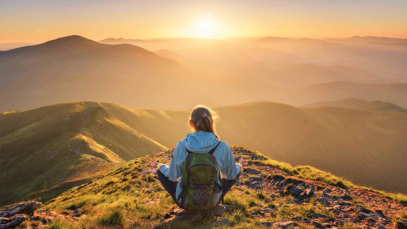 Kvinna med ryggsäck sitter och mediterar på ett berg i solnedgången